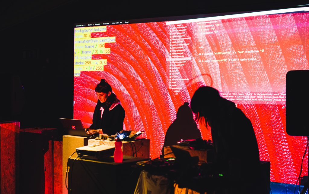 Two people on laptops in front of large projected red textures screen showing code