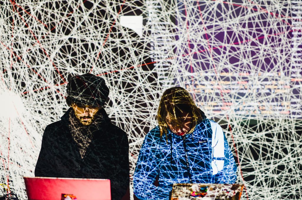 Two people on laptops in front of large projected screen showing live code and many overlapping lines