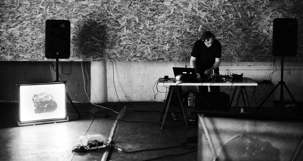 Black and white photo of man using laptop and other electrical equipment on a table with a projected image of a rock on a screen and large speakers set up