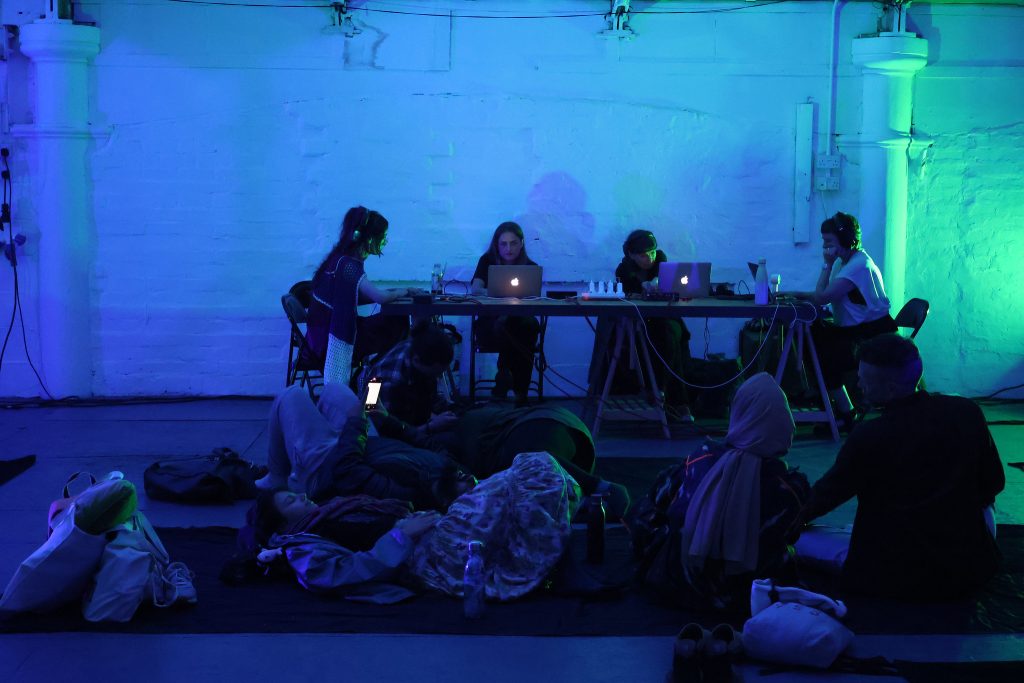 A group of people are making a sound performance under soft blue and green lights, they are focussed on their laptops an audience is watching them they are sitting on the floor