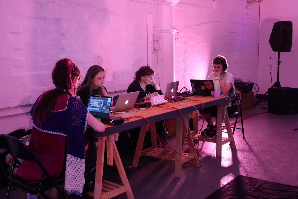 A group of people are making a sound performance under soft pink lights, they are focussed on their laptops