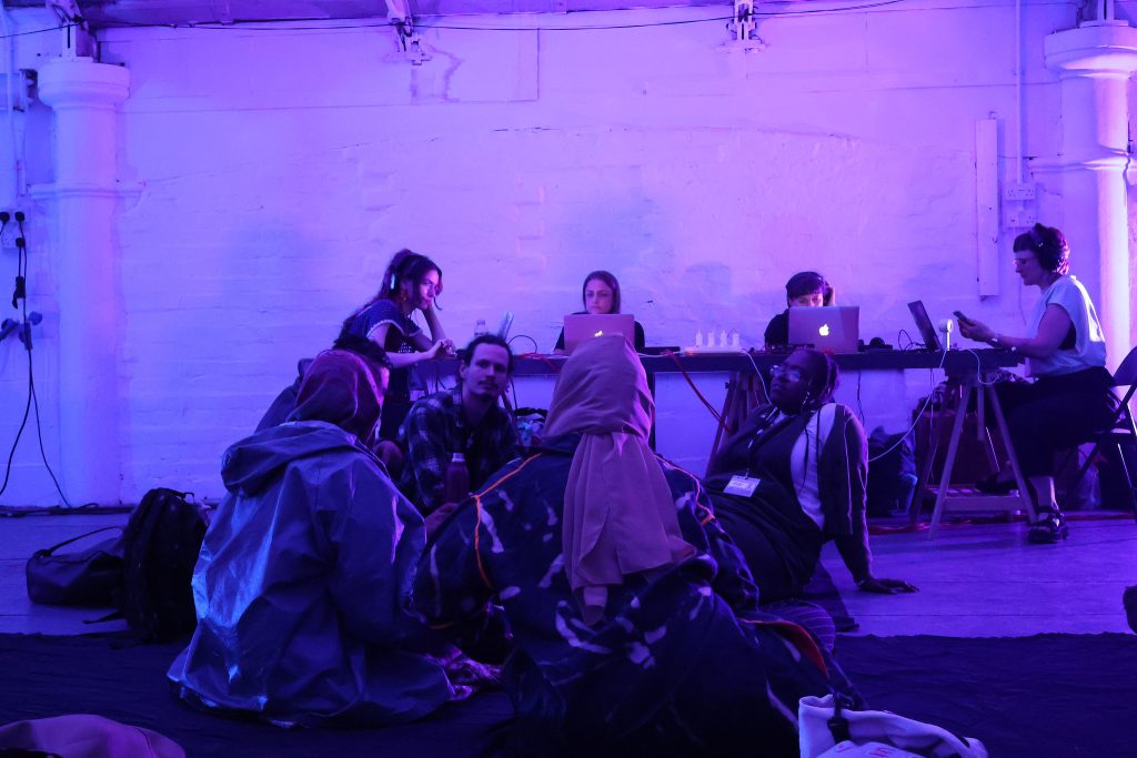 A group of people are making a sound performance under soft purple lights, they are focussed on their laptops an audience is watching them they are sitting on the floor