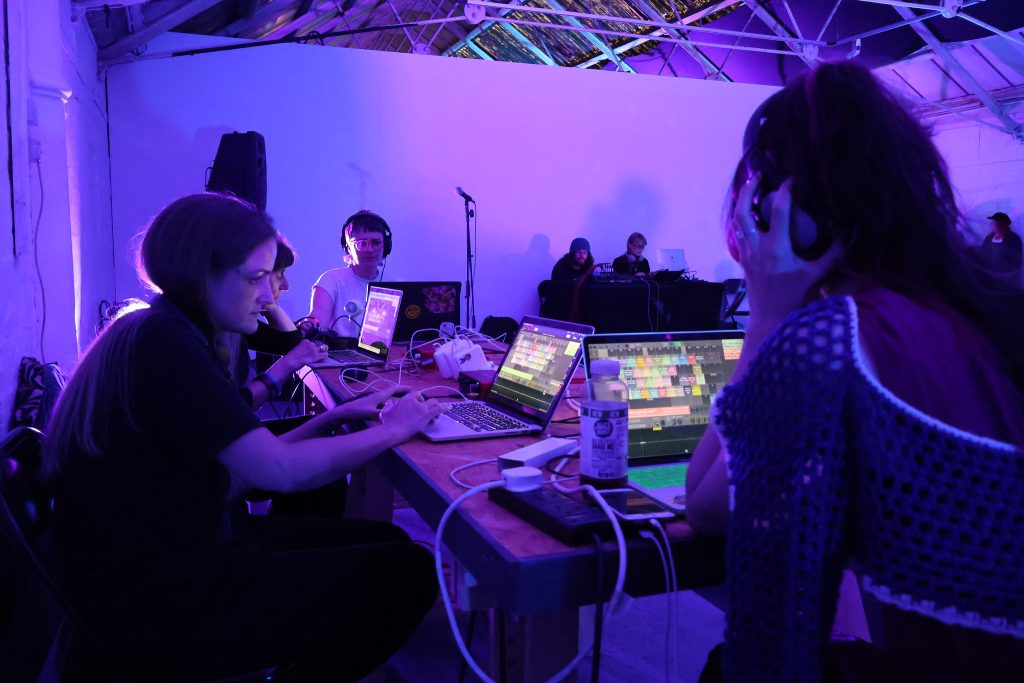 A group of people are making a sound performance under soft purple lights, they are focussed on their laptops
