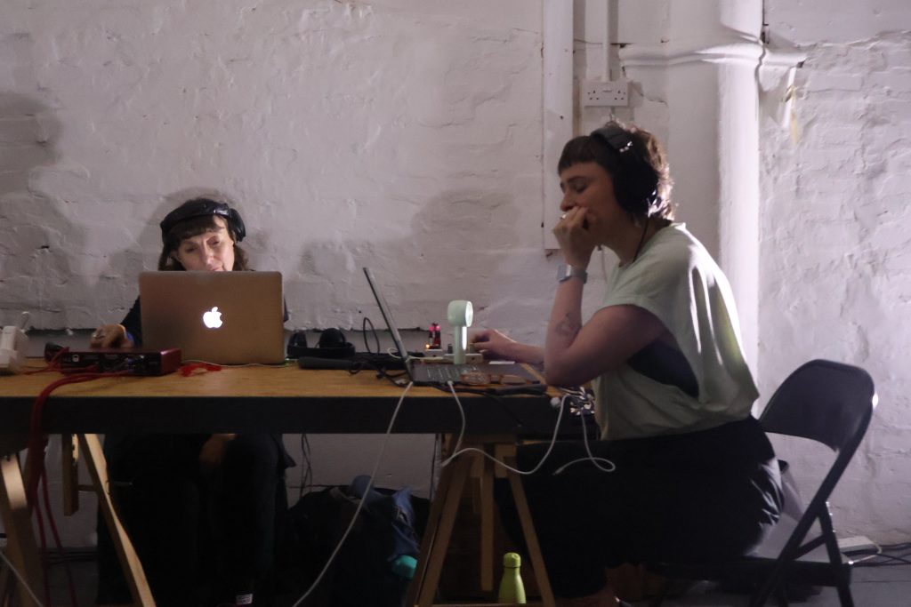 Two people are making a sound performance under soft white lights, they are focussed on their laptops