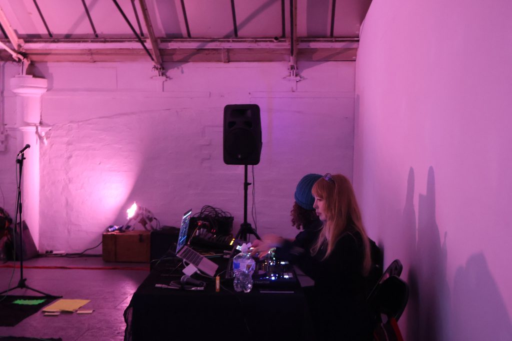 Two people are making a sound performance, they are sitting at a table focussed on what they are doing, the light is soft pink