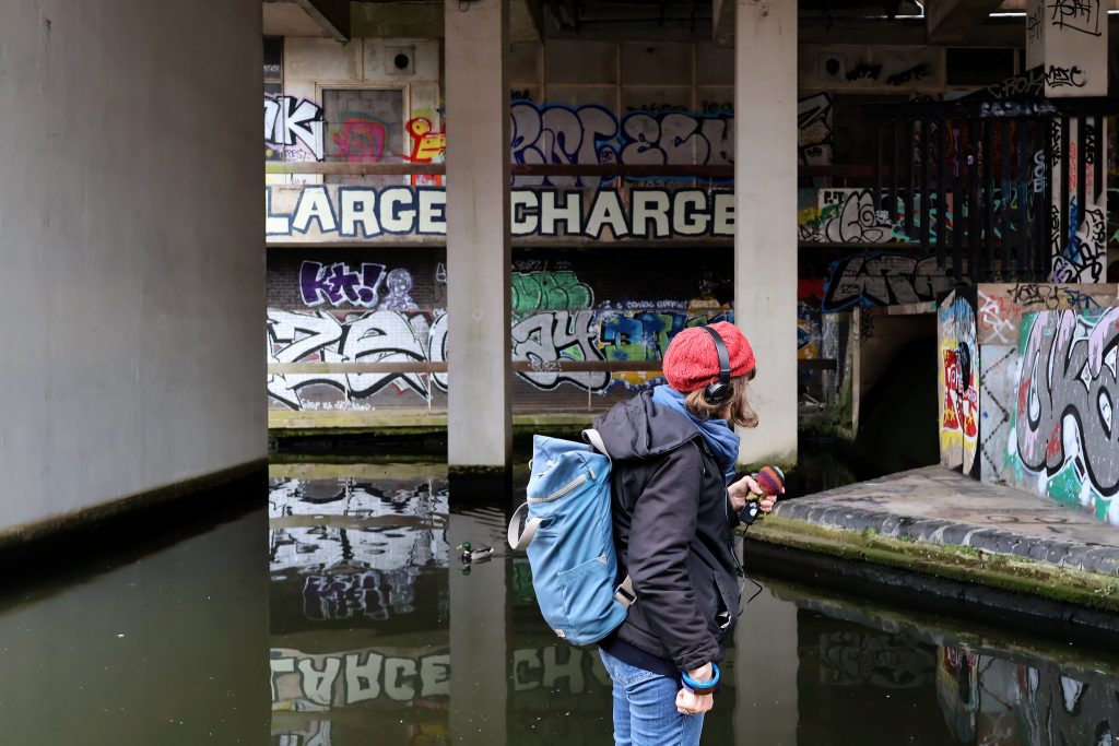 A woman in side profile is holding a sound recorder to make a field recording of the canal that runs next to the BT Tower in Birmingham