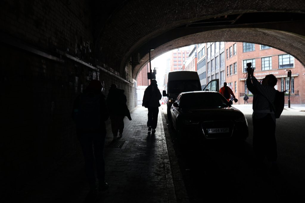 A group of people in silhouette are making a recording under a railway bridge on an industrial street in central Birmingham near Snow Hill Station