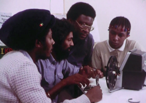 Four black men sat at a table around a film camera