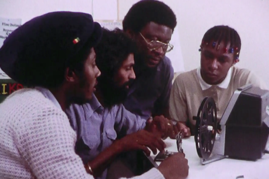 Four black men sat at a table around a film camera