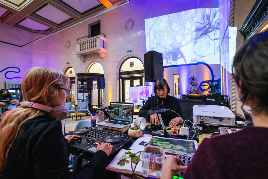 A laptop and music decks on a table alongside epemera being used by Cathy Wade to make visuals, which are projected onto the wall behind them