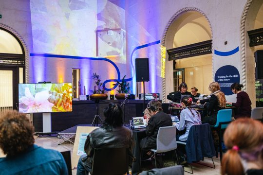the ways towards something collective sits around laptops at a table while visuals of flowers are projected onto a wall and shown on a monitor