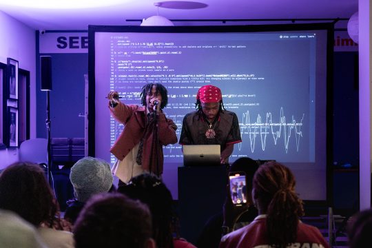Bundy on laptop and $witchblade performing with microphone and hand raised. A backdrop of code is behind then and the heads of some audience memebrs are visible