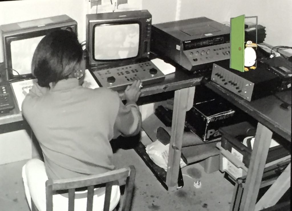 a black and white photographic image of a woman with her back to camera, seated at a video editing table. In the top right corner, a colourful animated penguin is walking out of a bright green door.