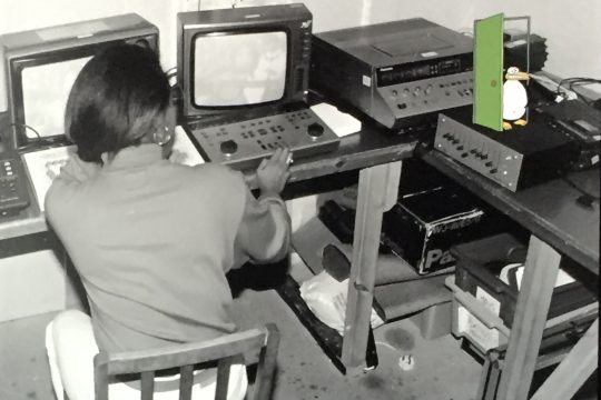 a black and white photographic image of a woman with her back to camera, seated at a video editing table. In the top right corner, a colourful animated penguin is walking out of a bright green door.