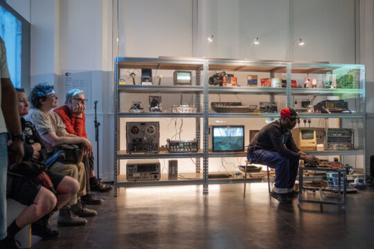 Gary Stewart in the Pixel Studio sat at a low table of electronic equipment, backed by shrlves full of analogue tech. An audience is seated to the left.