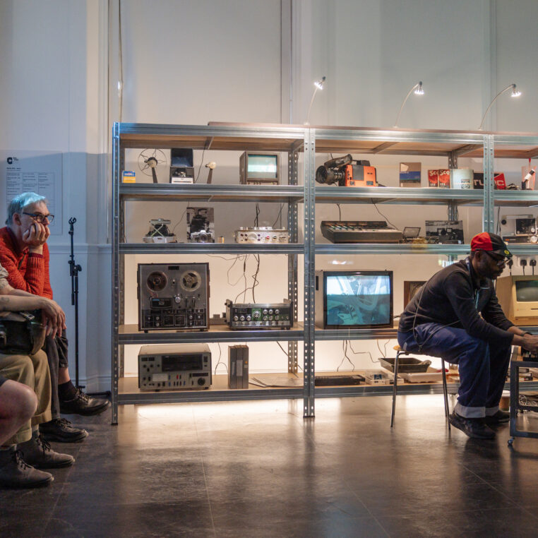 Gary Stewart in the Pixel Studio sat at a low table of electronic equipment, backed by shrlves full of analogue tech. An audience is seated to the left.