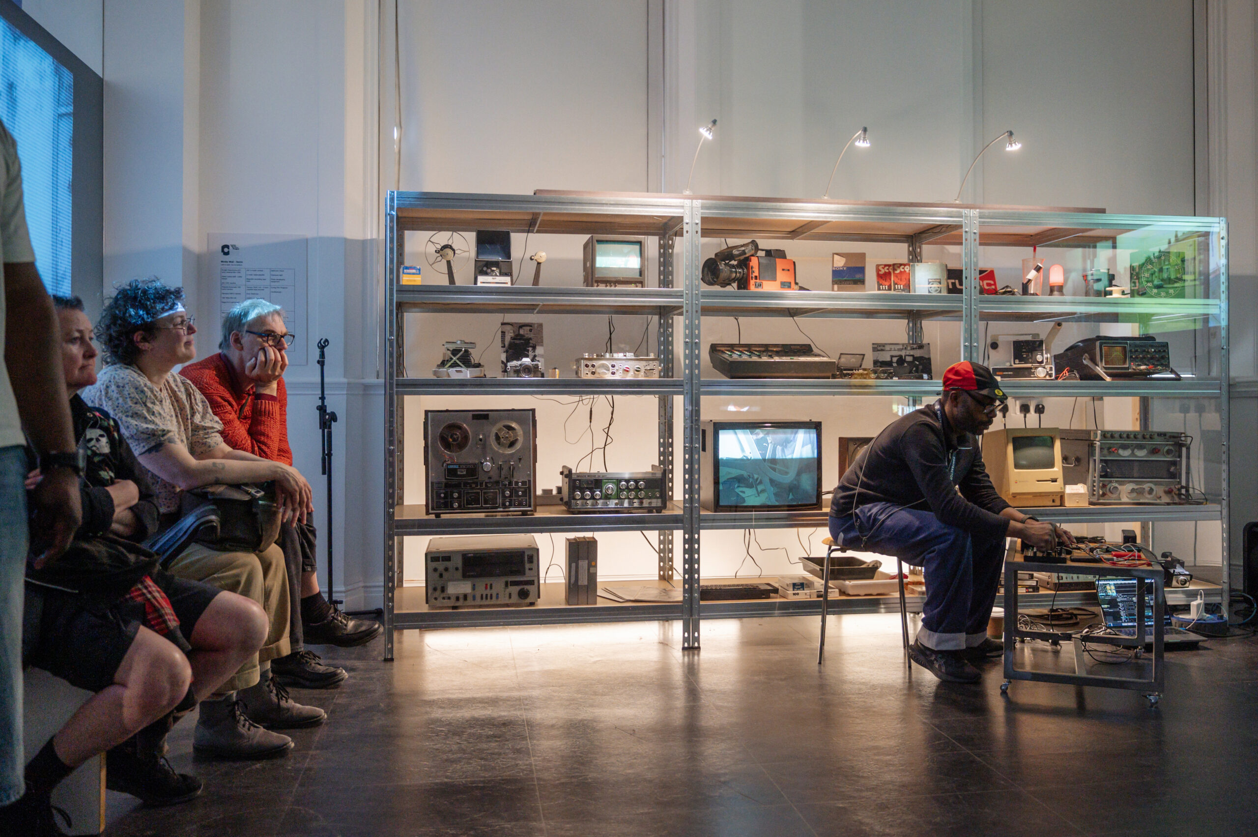 Gary Stewart in the Pixel Studio sat at a low table of electronic equipment, backed by shrlves full of analogue tech. An audience is seated to the left.